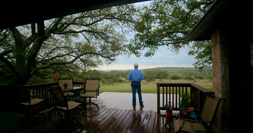 A person stands on a wooden deck, looking towards a vast green field with trees.