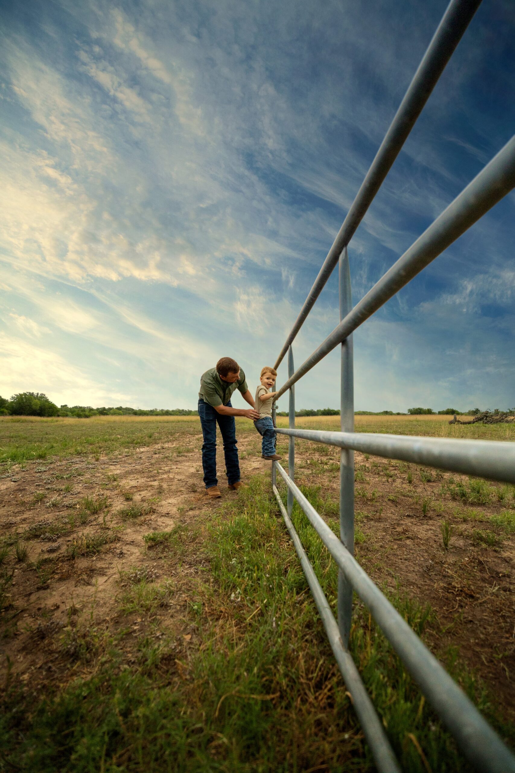 A father helping his kid climb a ranch fence.