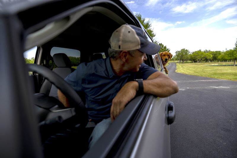 Man looking behind out of the driver seat window towards a dog