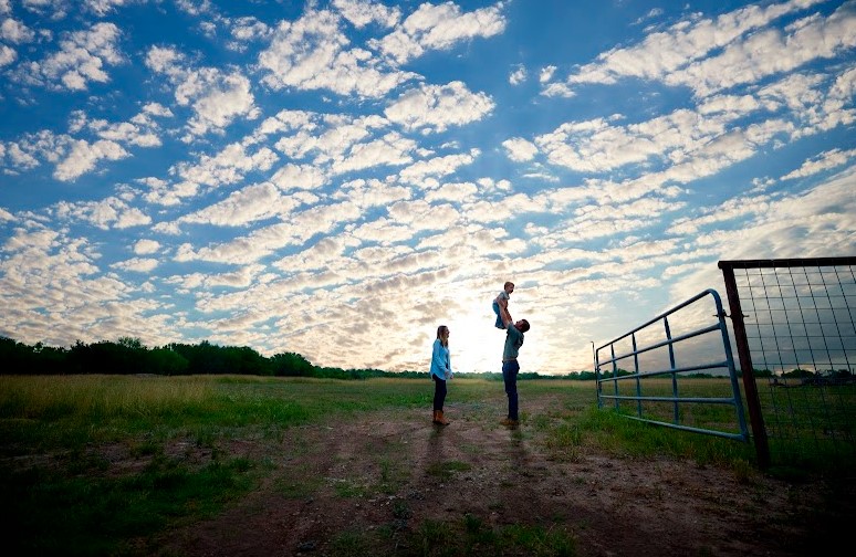 Three people, two adults and a child, stand in an open field near a metal gate under a blue sky with scattered clouds. One adult lifts the child overhead.