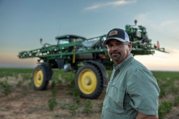 A man in a hat poses in front of a large tractor, surrounded by farmland.