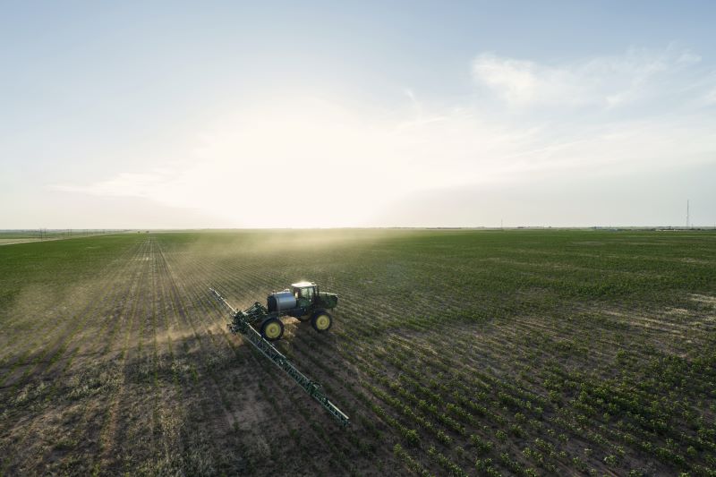 A tractor sprays crops in a large, flat agricultural field under a clear sky with the sun low on the horizon.
